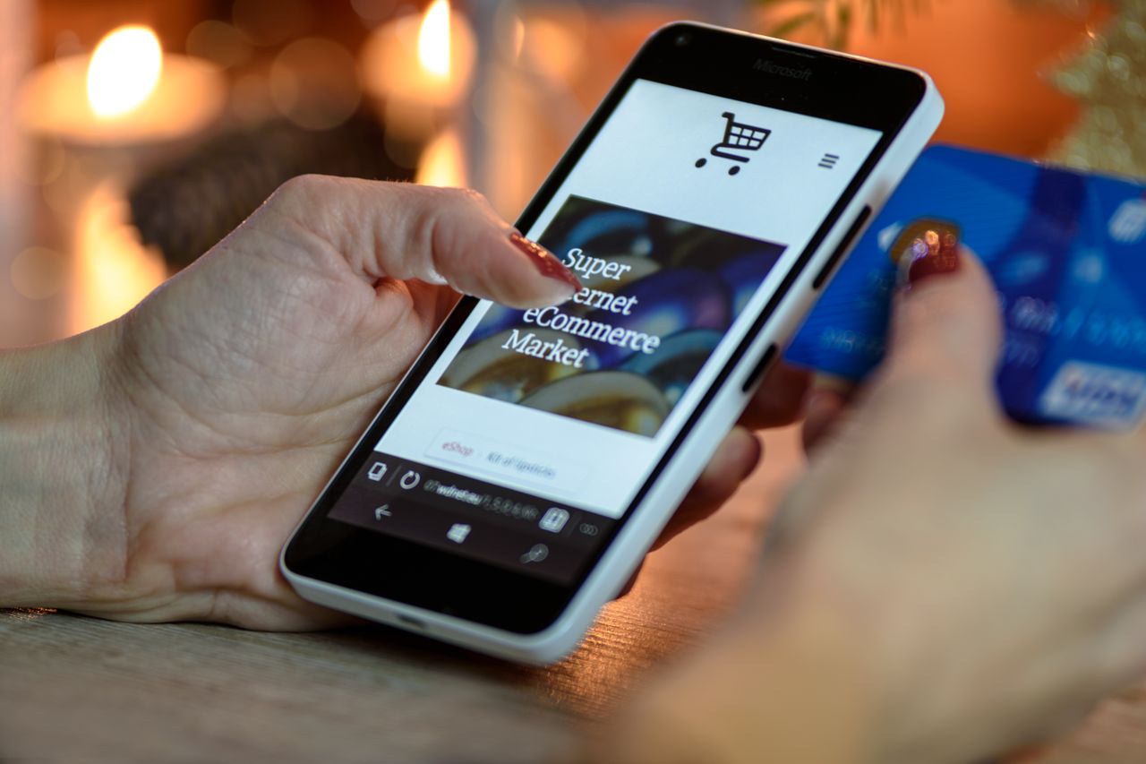 Femme utilisant un smartphone pour faire des achats en ligne avec une carte de crédit à la main, dans un décor aux lumières festives.